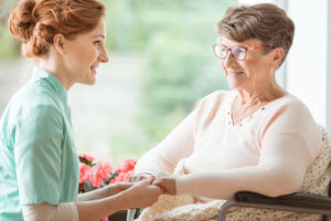 Compassionate CNA explaining a geriatric handicapped patient with dementia medical procedures while holding her hands. Assisted living house for pensioners. Side view.